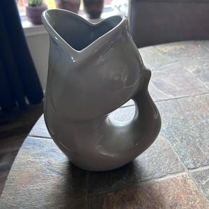 May include: A glossy, light gray ceramic pitcher shaped like a fish. The vessel features a wide, open mouth at the top that serves as a pouring spout, a curved body, and a hollow handle formed by the tail section of the fish design.