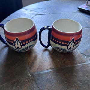 May include: Two ceramic mugs with a white interior, featuring a design of orange, burgundy, and blue bands with a floral motif. The mugs have dark blue handles and are set on a textured, dark gray surface.