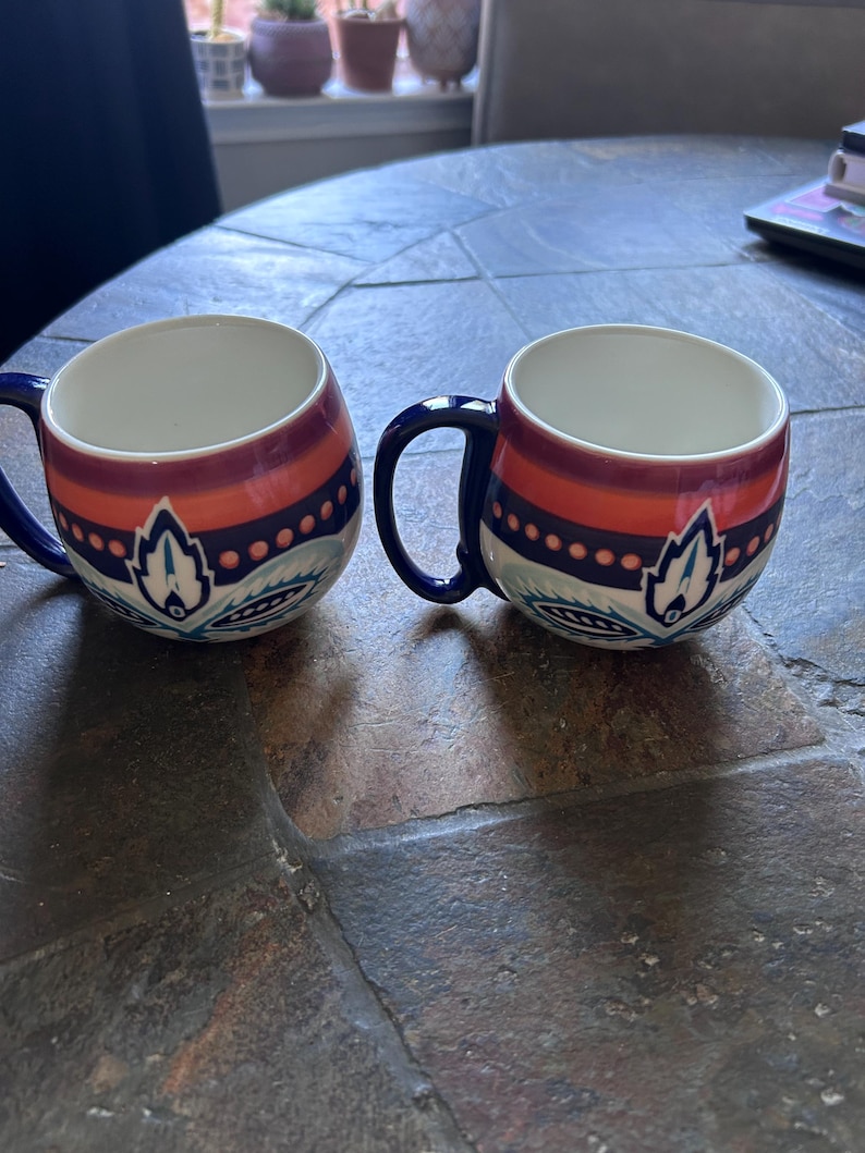 May include: Two ceramic mugs with a white interior, featuring a design of orange, burgundy, and blue bands with a floral motif. The mugs have dark blue handles and are set on a textured, dark gray surface.
