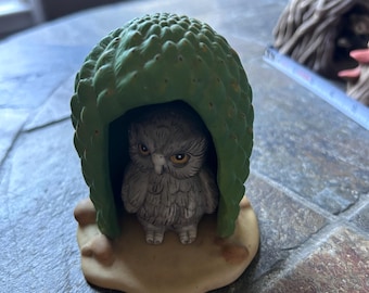 May include: A small ceramic figurine featuring a grey owl with yellow eyes standing inside a green, textured, dome-shaped structure that resembles a cactus or pinecone, all set upon a smooth, tan-colored base.