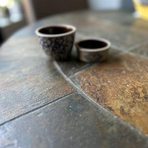 May include: Two small ceramic bowls with a dark interior and a blue floral pattern on a light background. The bowls are on a stone tile surface.