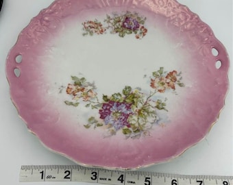 May include: A pink porcelain plate with a floral design. The plate has a scalloped edge and two small holes on either side. The plate is made in China and measures approximately 7 inches in diameter.