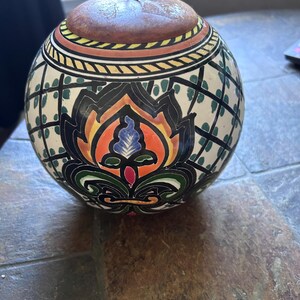 May include: A round, ceramic decorative jar with a lid. The jar features a hand-painted floral design in black, orange, blue, and green. The lid is brown with a metal ring handle. The jar is on a brown surface.