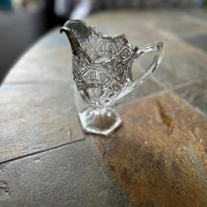 May include: A clear glass creamer with a decorative, geometric pattern. The creamer has a handle and a spout with a silver rim. It sits on a hexagonal base, showcasing a vintage design.