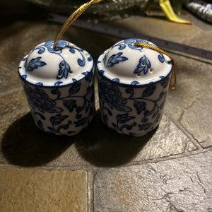 May include: Two matching ceramic salt and pepper shakers with a blue and white floral design. Each shaker has a rounded top with a gold-colored loop for hanging. The shakers are cylindrical and sit on a stone-like surface.