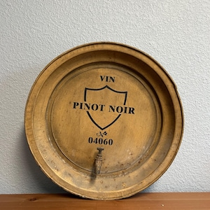 May include: Round, wooden wine barrel head with the text "VIN PINOT NOIR" in black. The barrel head has a spigot and the number "04060" below the text. The wood has a warm, brown tone with visible grain and a metal band around the edge.