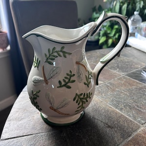 May include: White ceramic pitcher with a curved handle and spout. The pitcher is decorated with green leaves and dragonfly designs. The rim and base are trimmed with green. A decorative piece for serving beverages.