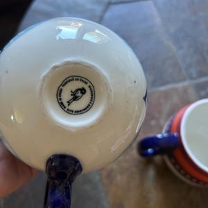 May include: White and blue ceramic teacup with a dark blue handle. The bottom of the cup has a circular stamp with text and a bird illustration. Another teacup with orange and blue stripes is in the background.