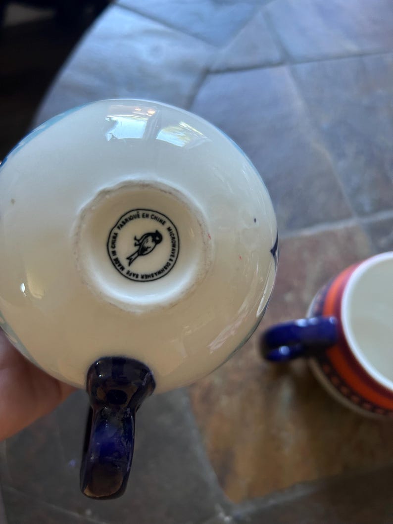May include: White and blue ceramic teacup with a dark blue handle. The bottom of the cup has a circular stamp with text and a bird illustration. Another teacup with orange and blue stripes is in the background.