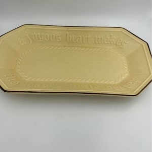 May include: A yellow rectangular serving dish with a brown rim. The dish has a raised design around the edge and the words "A joyous heart makes the weary taste better" are embossed on the surface.