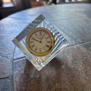 May include: A clear, diamond-shaped Waterford crystal desk clock featuring a gold-tone bezel and a white face with black Roman numerals. The clock face displays the text Waterford Crystal in black lettering, with black hour, minute, and second hands.