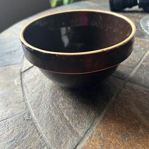 May include: A dark brown ceramic bowl with a light tan rim. The bowl has a wide, rounded shape and sits on a stone surface. The interior of the bowl is a glossy dark brown.