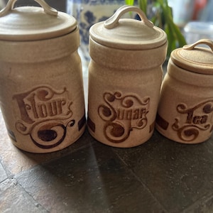 May include: Three beige ceramic canisters with lids and handles, labeled "Flour", "Sugar", and "Tea" in a decorative font. The canisters have a rustic, textured appearance and are arranged on a dark surface.
