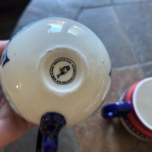 May include: White ceramic mug with blue star pattern and a dark blue handle. The bottom of the mug has a stamp that reads "Fabriqu&eacute; en Chine Microwave & Dishwasher Safe". Another mug with a red and blue striped design is visible.