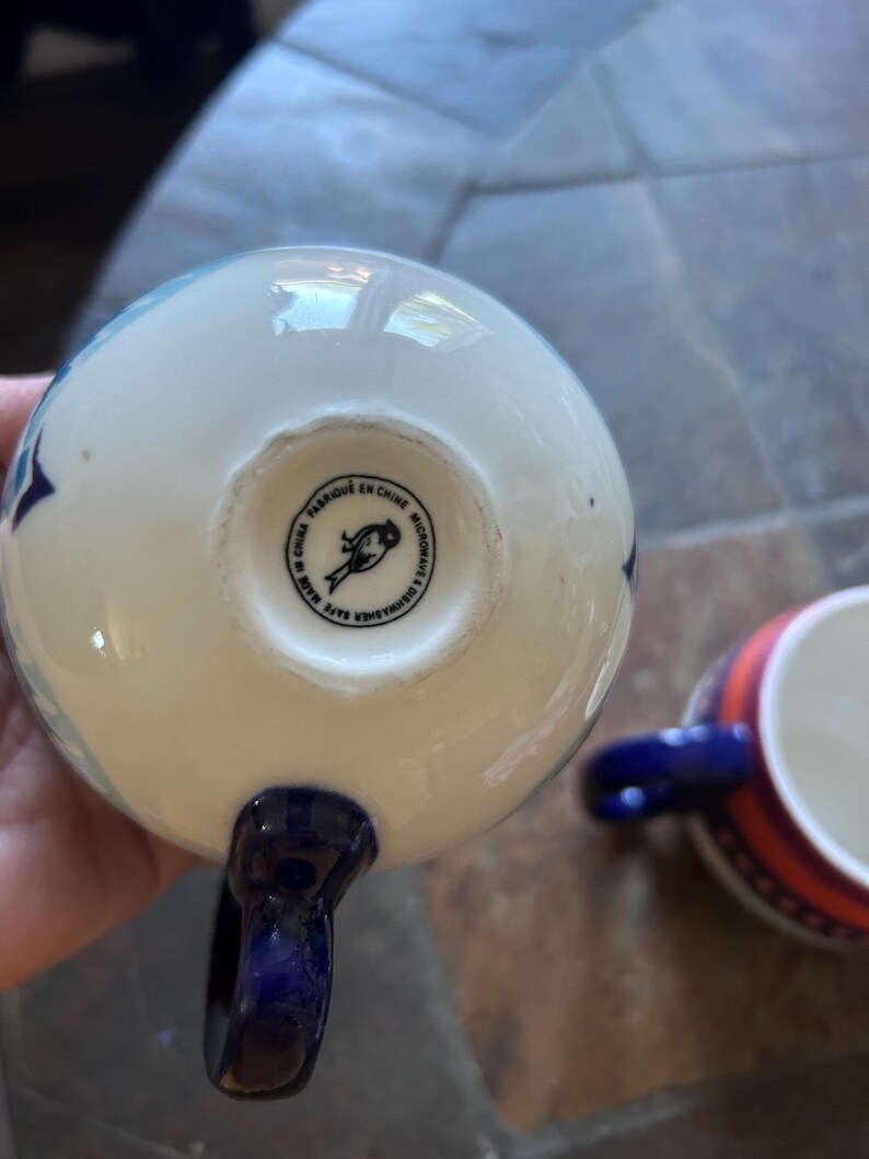May include: White ceramic mug with blue star pattern and a dark blue handle. The bottom of the mug has a stamp that reads "Fabriqu&eacute; en Chine Microwave & Dishwasher Safe". Another mug with a red and blue striped design is visible.