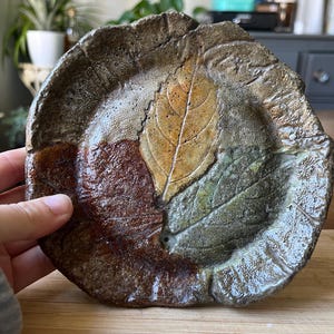 May include: Handmade ceramic plate with a leaf design. The plate features a textured surface with a brown, green, and tan color scheme. The leaf imprints are detailed, adding a natural aesthetic. A unique piece for serving or display.
