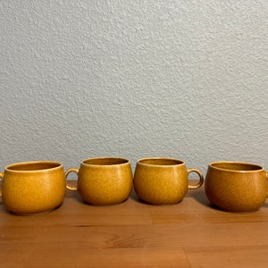 May include: Four brown ceramic mugs with rounded sides and handles. The mugs are arranged in a row on a wooden surface.