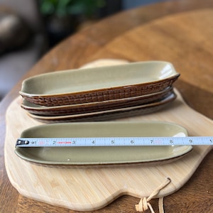 May include: A set of four brown and beige ceramic serving dishes stacked on a wooden cutting board. The dishes are rectangular and have a textured surface. A tape measure is placed on top of the dishes, showing a measurement of 9 inches.