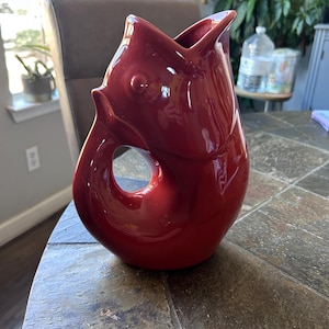 May include: A glossy, red ceramic pitcher in the shape of a fish. The pitcher has a large opening at the top and a circular opening near the base, resembling a fish's mouth and eye. The design is a unique decorative item.
