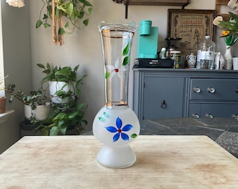 May include: A tall, clear glass vase with a frosted base and a flared top. The vase is decorated with hand-painted white and blue flowers with green leaves. The vase is sitting on a wooden surface.
