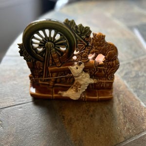 May include: A ceramic figurine depicting a dog and cat interacting with a spinning wheel. The figurine is primarily brown with green and white accents. The dog is white with brown spots and the cat is brown. The wheel is green.