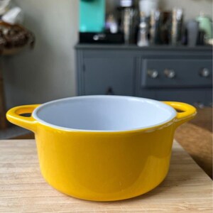 May include: A small yellow ceramic ramekin with white interior and two handles. The ramekin is sitting on a wooden cutting board.