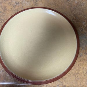 May include: A round, ceramic plate with a light beige center and a dark brown rim. The plate is set against a mottled brown surface. This plate is suitable for serving food.