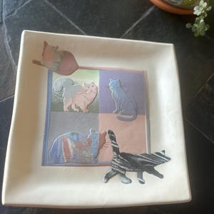 May include: Square ceramic dish with embossed cat designs in various colors. The dish features a cream-colored border and a central square divided into four sections, each with a cat illustration. The cats are in shades of pink, blue, and gray.