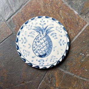 May include: A round, white ceramic coaster with a hand-painted blue pineapple design in the center. The coaster has a blue border with a repeating leaf pattern. The coaster is approximately 4 inches in diameter.