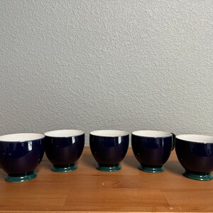 May include: Five dark blue teacups with a green rim, arranged in a row on a wooden surface.