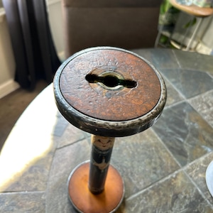 May include: A vintage wooden spool with a metal rim and base. The spool has a keyhole-shaped opening in the center. The wooden parts are a warm brown color, and the metal is a dark gray. The spool is standing upright.