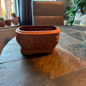 May include: A terracotta-colored rectangular planter with a detailed relief design of figures. The planter has a dark red interior and sits on a dark, textured surface. The planter is approximately 12 inches long.
