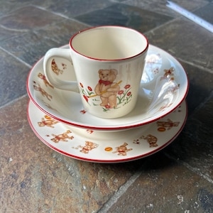 May include: A cream-colored children's tea set with a teddy bear theme. The set includes a small mug, a bowl, and a plate, all with a red trim. The mug, bowl, and plate are decorated with images of teddy bears and flowers.