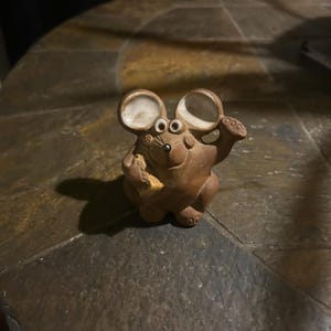 May include: A brown ceramic mouse figurine with large ears, round eyes, and a small nose. The mouse is holding a piece of cheese. The figurine is on a stone surface.