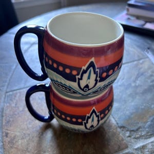 May include: Two stacked ceramic mugs with a white interior and a colorful exterior design. The mugs feature a dark blue handle and a pattern of orange, purple, and blue stripes with a stylized leaf design. The mugs are on a stone surface.
