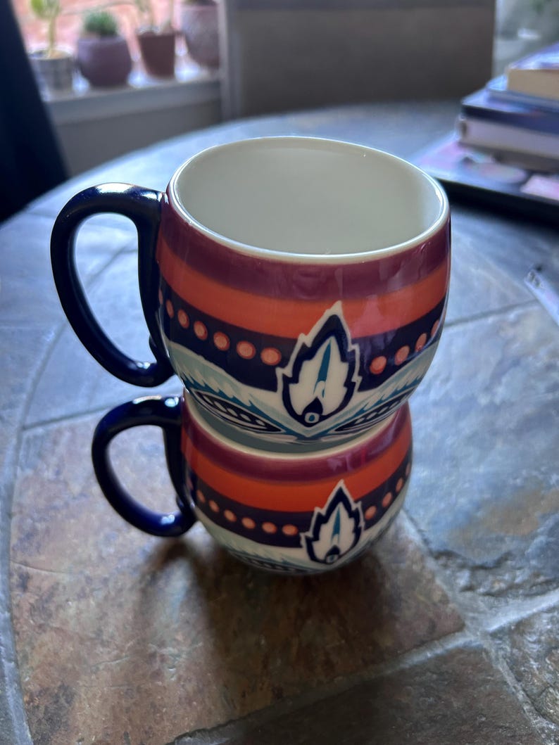 May include: Two stacked ceramic mugs with a white interior and a colorful exterior design. The mugs feature a dark blue handle and a pattern of orange, purple, and blue stripes with a stylized leaf design. The mugs are on a stone surface.