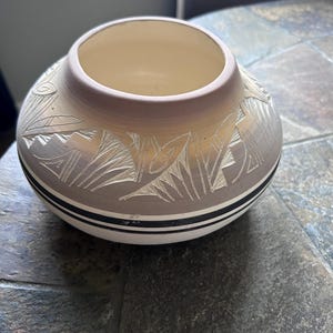 May include: A round ceramic bowl with a wide opening, featuring a cream-colored interior and a light tan exterior. The exterior is decorated with carved geometric patterns and two thin black horizontal lines encircling the lower section.