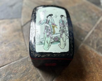 May include: Black rectangular box with a decorative lid featuring a painted scene of figures in traditional attire. The box has a black exterior with a detailed border and a glossy finish. The lid's artwork includes pastel colors and intricate details.