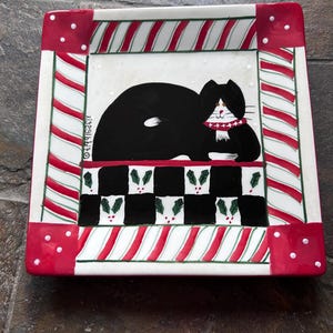 May include: A square ceramic plate featuring a black and white cat wearing a red scarf. The center design includes a black and white checkered pattern with holly leaves. The border has red and white candy cane stripes and red corners.
