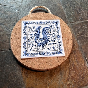 May include: A square, white ceramic tile featuring a blue rooster design, framed by blue floral and leaf motifs. The tile rests on a round cork trivet with a rope handle. The rooster is a traditional design.