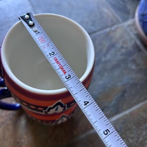 May include: A ceramic mug with a colorful design, featuring orange, red, and blue accents. The mug is approximately 3 inches (7.6 cm) in diameter, as measured by a tape measure. The mug has a blue handle.