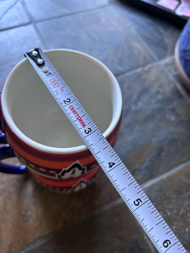 May include: A ceramic mug with a colorful design, featuring orange, red, and blue accents. The mug is approximately 3 inches (7.6 cm) in diameter, as measured by a tape measure. The mug has a blue handle.