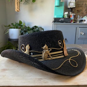 May include: Black straw cowboy hat with decorative gold-colored cat figurines and accents. The hat features a wide brim and a structured crown. The cat figurines are adorned with rhinestones. The hat is displayed on a wooden surface.