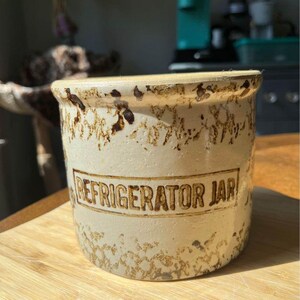 May include: Cream-colored vintage refrigerator jar with brown speckled detailing. The jar has a cylindrical shape with a lid and the words "REFRIGERATOR JAR" are printed on the front. The jar is sitting on a wooden surface.