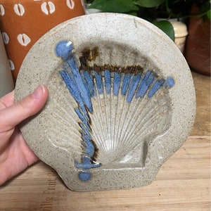 May include: A ceramic dish shaped like a seashell with a blue and brown glaze. The dish has a textured surface and a raised rim.