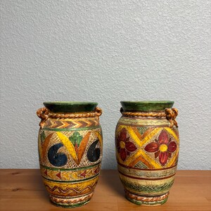 May include: Two decorative ceramic vases with a colorful, patterned design. The vases have a green rim, rope-like handles, and a base with a repeating geometric pattern. One vase features a floral design, while the other has a more abstract pattern.
