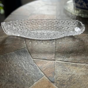 May include: A clear, oval-shaped glass dish with a detailed floral pattern around the rim and a textured surface. The dish is empty and sits on a stone-like surface, reflecting light.