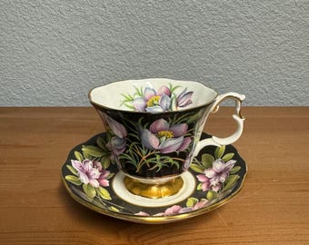 May include: A black teacup and saucer with a floral design. The cup and saucer feature a black background with pink and purple flowers and green leaves. The cup has a gold rim and a gold base. The saucer has a gold rim.