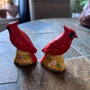May include: Two red cardinal figurines perched on decorative bases. The bases are tan with floral accents and the words "North Carolina" in black. The cardinals have black accents around their eyes and beaks.