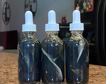 May include: Three clear glass dropper bottles filled with a dark liquid and plant matter. Each bottle has a white dropper top. The bottles are arranged in a row on a brown countertop.